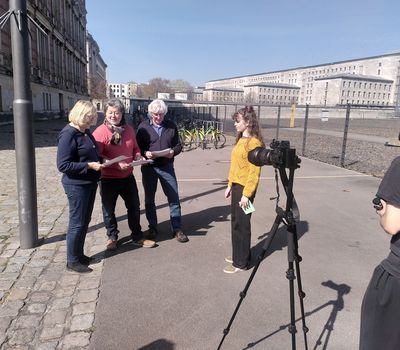 Drei ältere Erwachsenen steht eine junge Frau gegenüber, während eine weitere Person die Szene mit einer Kamera auf einem Stativ filmt. Sie stehen auf einer gepflasterten Fläche im Freien. Im Hintergrund große Steinbauten und ein eingezäunter Bereich.