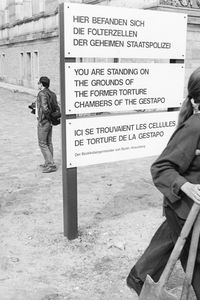 Eine Frau und ein Mann mit Spaten gehen an einem Schild vorbei mit der Aufschrift: „Hier befanden sich die Folterzellen der Gestapo“, mit Übersetzungen auf Englisch und Französisch.