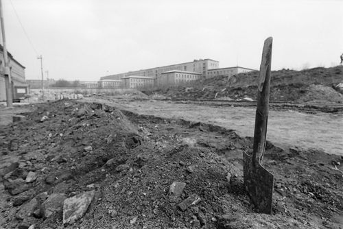Im Vordergrund steckt ein Spaten mit durchgebrochenem Stiel in einem Haufen aus Aushub, der sich in gerader Linie fortsetzt. Dahinter eine leere Fläche mit Reifenspuren. Im Hintergrund ein mit Gras bewachsener Erdhügel und ein massives Gebäude.