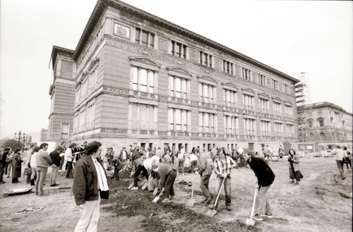 Menschen graben mit Spaten auf einem offenen Gelände vor einem historischen Gebäude mit stuckverzierter Fassade. Eine Seite des rechtwinkligen Grabens verläuft parallel zur Gebäudefront. Links und rechts stehen weitere Beteiligte und Zuschauende.