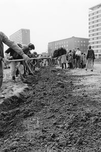 Mehrere Menschen graben in einer langen Reihe mit Spaten einen flachen Graben in aufgewühlter Erde. Rechts moderne mehrstöckige Gebäude. Im Hintergrund einige zuschauende Personen.