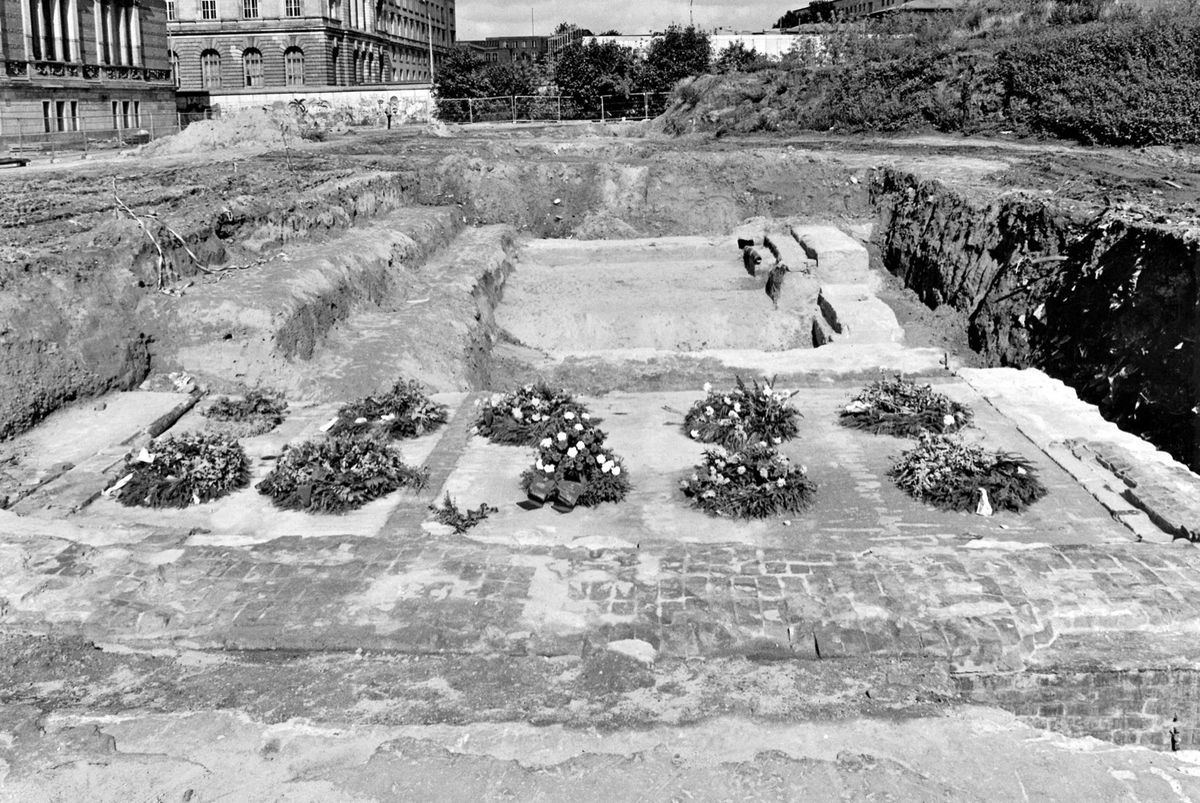 Schwarz-weiß-Fotografie: Blick auf eine Ausgrabungsstätte mit freigelegtem Mauerwerk und gepflastertem Boden. Mehrere Blumenkränze sind auf dem Fundament angeordnet. Im Hintergrund ein historisches Gebäude sowie unbebautes Gelände mit Erdaufschüttungen.