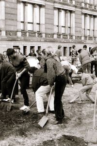 Eine Gruppe von Menschen, darunter auch ein Kind, steht vor einem historischen Gebäude und gräbt nebeneinander mit Spaten in dunkler Erde. Einige beugen sich vor, andere schauen pausierend auf. Im Hintergrund ein Bauwagen und beobachtende Personen.