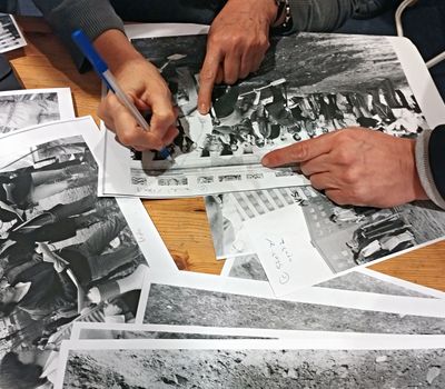 Auf einem Tisch liegen viele Ausdrucke historischer Fotografien. Eine Hand schreibt auf ein Schwarz-Weiß-Foto, zwei weitere Hände deuten auf verschiedene Stellen.