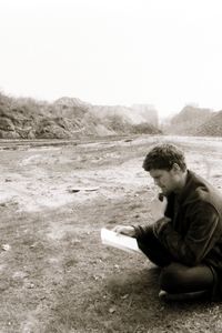 Schwarz-weiß-Fotografie. Ein Mann sitzt im Schneidersitz auf dem Boden einer flachen Brache und liest, eine Hand ans Kinn gelegt, einen Zettel. In der Ferne ein mit Gras bewachsener Erdhügel, daneben ein Kiesberg. Auf dem Erdhügel steht eine Person.