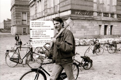 Mann mit Spaten über der Schulter, der vor einem historischen Gebäude geht. Im Hintergrund ein Schild mit der Aufschrift: „Hier befanden sich die Folterzellen der Geheimen Staatspolizei.“, darunter eine englische und französische Übersetzung.