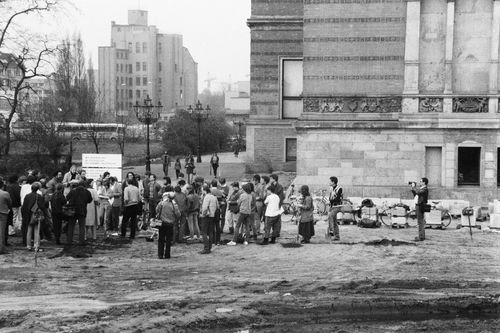 Eine Menschengruppe steht auf einem unbefestigten Gelände neben einem historischen Gebäude. Im Vordergrund Reifenspuren, rechts hält eine Person eine Kamera und fotografiert die Gruppe. Im Hintergrund Bäume und moderne Gebäude.