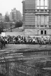 Etwa 80 Menschen graben auf offenem Gelände vor einem großen Gebäude. Die Grabung ist L-förmig, die lange Seite verläuft parallel zur Gebäudefront. Im Vordergrund Reifenspuren und eine Pfütze.