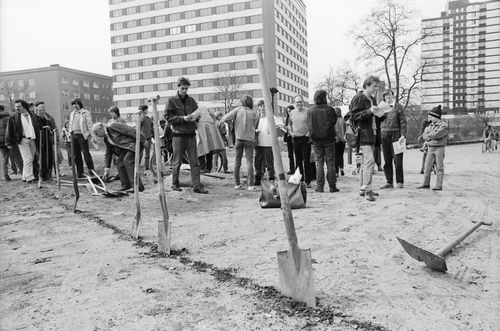 Entlang einer in die Erde gezogenen Linie stecken Spaten. Rechts liegt eine Schaufel auf dem Boden. Dahinter stehen mehrere Personen auf einem sandigen Gelände zwischen Wohnhäusern. Einige lesen von Zetteln oder unterhalten sich.