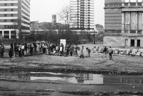 Menschen stehen und graben auf einem unbefestigten Gelände neben einem historischen Gebäude. Im Vordergrund eine große Pfütze mit Reifenspuren, im Hintergrund zwei moderne mehrstöckige Gebäude und Bäume.