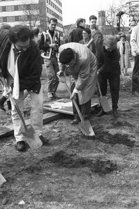 Vier Personen beginnen entlang einer in die Erde geritzten Markierung zu graben. Im Hintergrund stehen Menschen und beobachten das Geschehen. Drei von ihnen halten Zettel in der Hand und schauen aufmerksam zu.