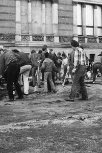 Zahlreiche Menschen graben mit Spaten auf einem unbefestigten Gelände neben einem historischen Gebäude. Sie arbeiten in L-Form, die lange Seite verläuft entlang der Gebäudefront. Links kniet eine Person mit Kamera, im Vordergrund harter Boden.