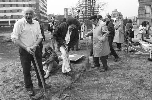 Auf einem offenen Gelände stehen verstreut Menschen. Im Vordergrund ist eine Linie in die Erde geritzt, an der Menschen mit Spaten stehen. Ein älterer Mann blickt in die Kamera, ein Mann beginnt zu graben, ein dritter blickt auf die Markierung.