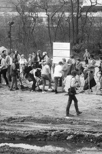 Zahlreiche Menschen versammeln sich auf einem unbefestigten Gelände. Einige stehen, andere graben mit Spaten. Im Vordergrund eine große Pfütze, im Hintergrund Bäume und moderne Gebäude.