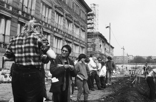 Vor einem historischen Gebäude stehen Menschen am Rand eines aufgewühlten Erdabschnitts. Im Vordergrund dreht ein Mann eine Zigarette. Rechts stehen Spaten und Schaufeln in den umgegrabenen Erdabschnitt gesteckt.