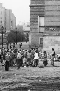 Eine Menschengruppe steht auf einem unbefestigten Gelände neben einem historischen Gebäude. Im Vordergrund Reifenspuren, rechts hält eine Person eine Kamera und fotografiert die Gruppe. Im Hintergrund Bäume und moderne Gebäude.