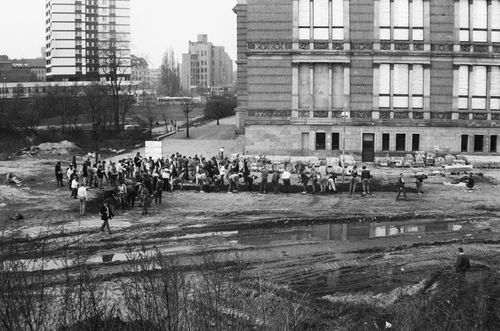 Etwa 80 Menschen graben auf offenem Gelände vor einem großen Gebäude. Die Grabung ist L-förmig, die lange Seite verläuft parallel zur Gebäudefront. Im Vordergrund Reifenspuren und eine Pfütze.