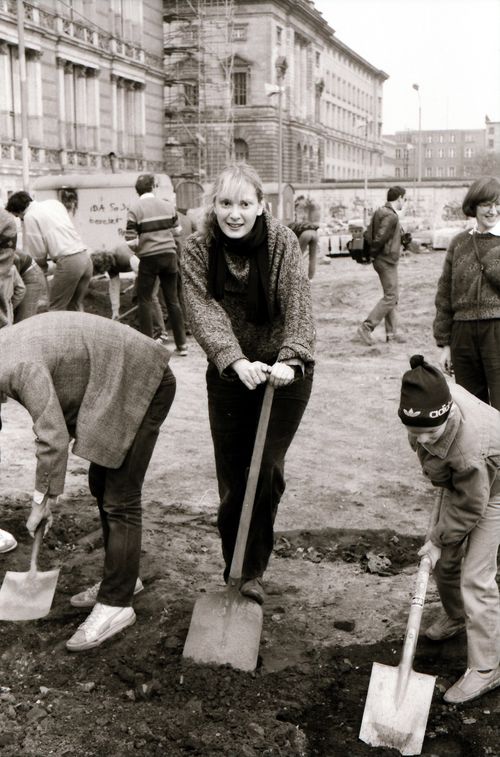 Eine Frau blickt freundlich in die Kamera und stützt einen Fuß auf einen Spaten. Neben ihr graben zwei Personen in der Erde, darunter ein Kind mit Mütze. Im Hintergrund Baugerüste und historische Gebäude.
