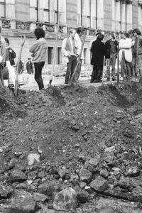 Ein Erdhaufen markiert den Verlauf eines Grabens, dessen lange Seite parallel zu einem historischen Gebäude verläuft. Die kurze Seite liegt im Vordergrund des Bildes. Entlang des Haufens stecken Spaten und Schaufeln, im Hintergrund stehen Menschen.