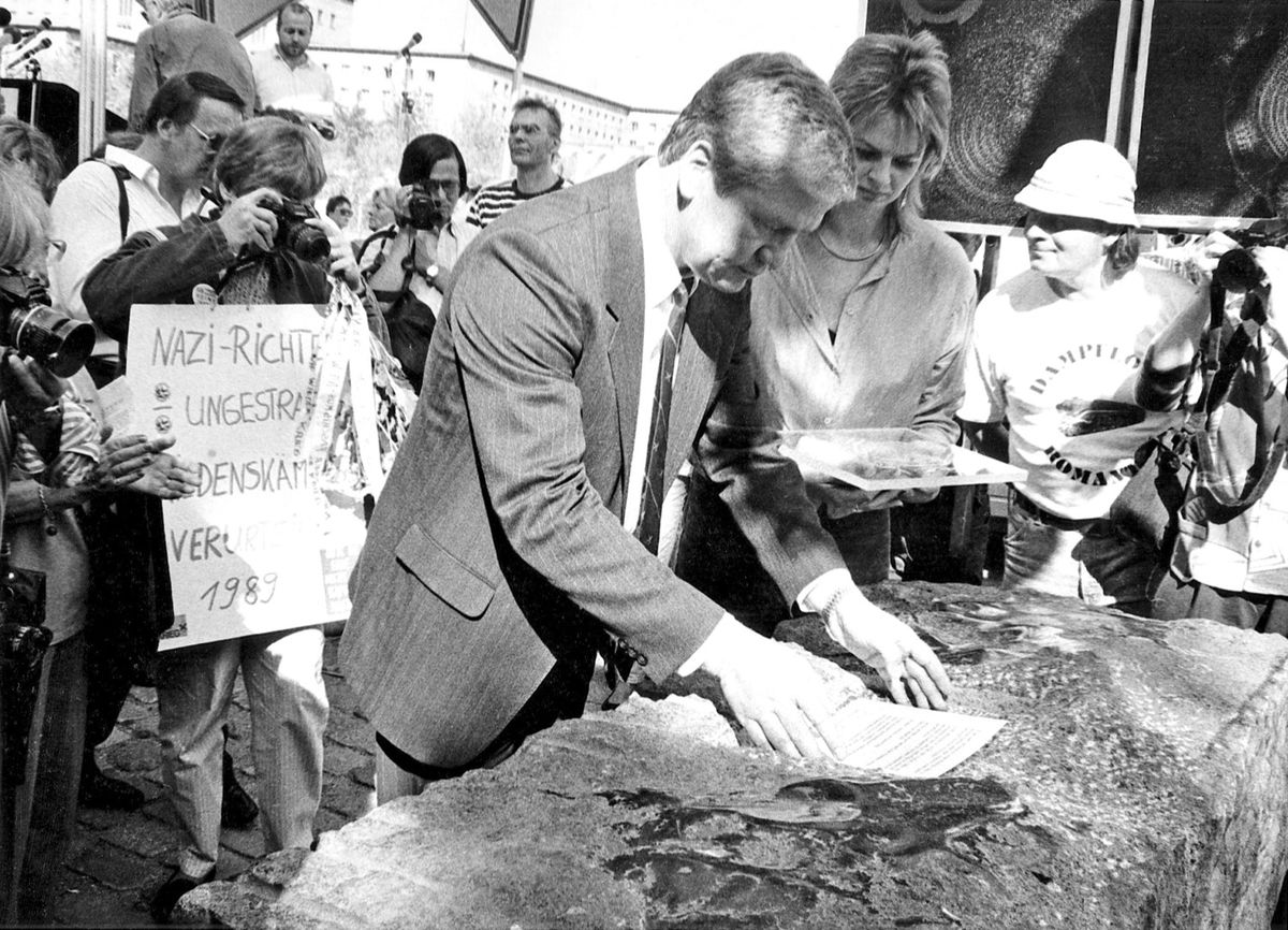 Ein Mann bringt eine Inschrift auf einem Steinblock an, umgeben von weiteren Personen. Links halten Demonstrierende ein Schild mit der Aufschrift „Nazi-Richter ungestraft – Gedenkstätte verhindert 1989“. Mehrere Fotograf*innen dokumentieren die Szene.