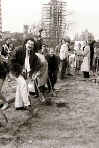 Eine Gruppe von Menschen steht auf einem brachliegenden Gelände. Viele von ihnen halten Spaten in der Hand und beginnen gemeinsam zu graben. In der Erde ist als Markierung eine gerade Linie geritzt.