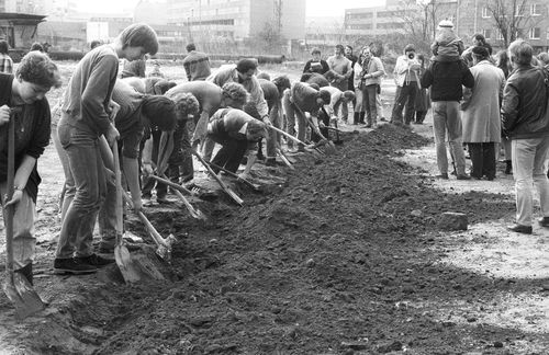 Entlang eines langen aufgeworfenen Erdwalls graben zahlreiche Personen dicht nebeneinander mit Spaten. Rechts steht eine größere Gruppe von Zuschauenden, darunter ein Kind auf den Schultern eines Erwachsenen. Im Hintergrund moderne Gebäude.