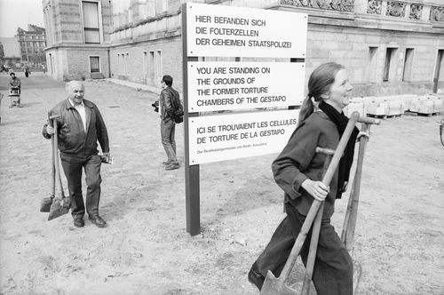 Eine Frau und ein Mann mit Spaten gehen an einem Schild vorbei mit der Aufschrift: „Hier befanden sich die Folterzellen der Gestapo“, mit Übersetzungen auf Englisch und Französisch.