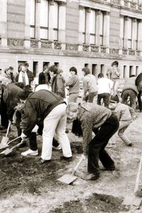 Eine Gruppe von Menschen, dicht nebeneinander stehend, gräbt mit Spaten eine L-förmige Rinne in dunkler Erde vor einem historischen Gebäude. In der Mitte des rechtwinkligen Grabens steht ein Kind mit Spaten. Im Hintergrund ein Bauwagen und Zuschauende.