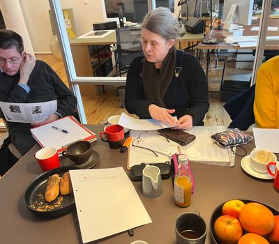 Drei Personen sitzen an einem Tisch, der mit Papieren, Stiften, Obst und Kaffeetassen bedeckt ist. Sie wirken konzentriert und sind in Diskussion oder Lektüre vertieft. Der Raum ist hell, mit Glaswänden und Holzboden gestaltet.