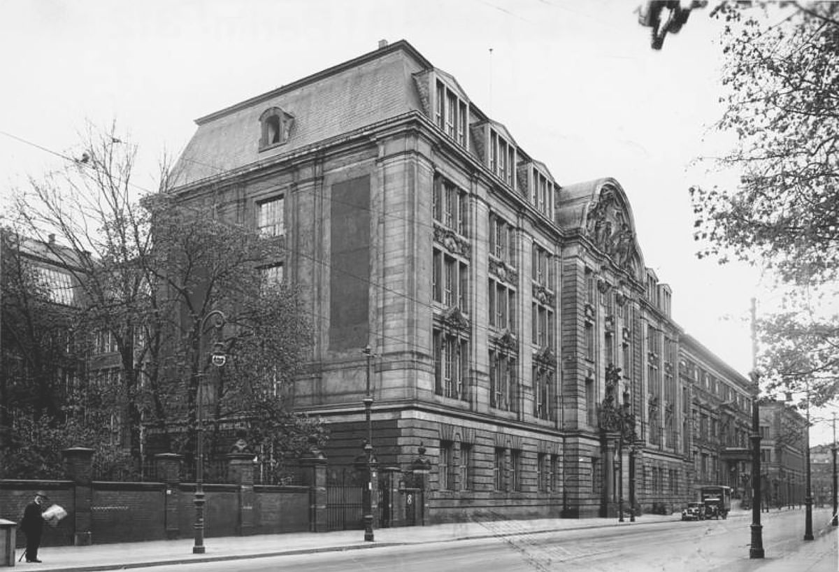 Schwarz-weiß-Fotografie: Monumentales, fünfstöckiges Gebäude mit reich verzierter Fassade und Mansarddach. Davor eine ruhige Straße mit Gehweg, Straßenlaternen und einem Passanten mit Zeitung.