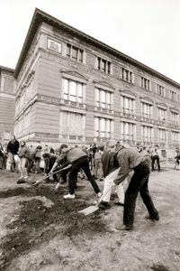 Mehrere Personen graben mit Spaten in dunkler Erde. Die Grabung hat gerade begonnen. Im Hintergrund ein mit Stuck verziertes, historisches Gebäude.